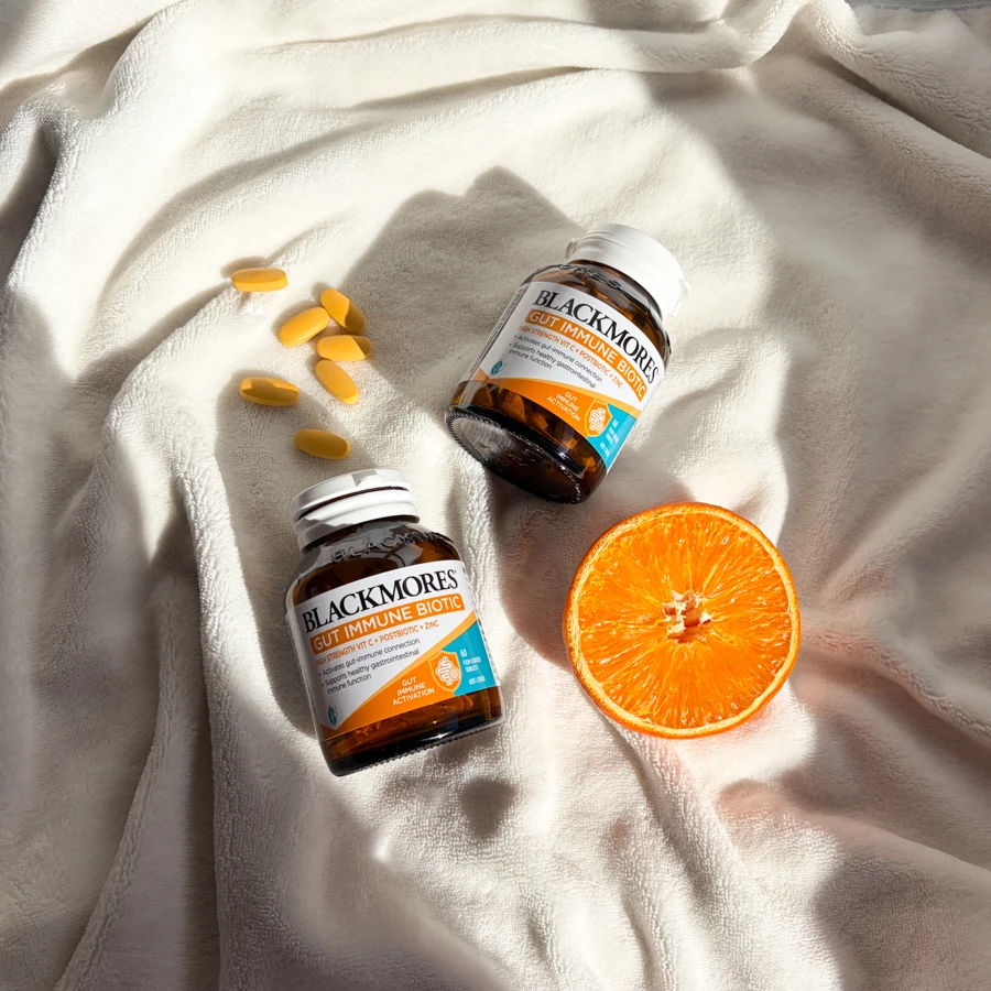 Two Blackmores Gut Immune Biotic supplement bottles, yellow tablets, and a halved orange on a cream fabric surface.