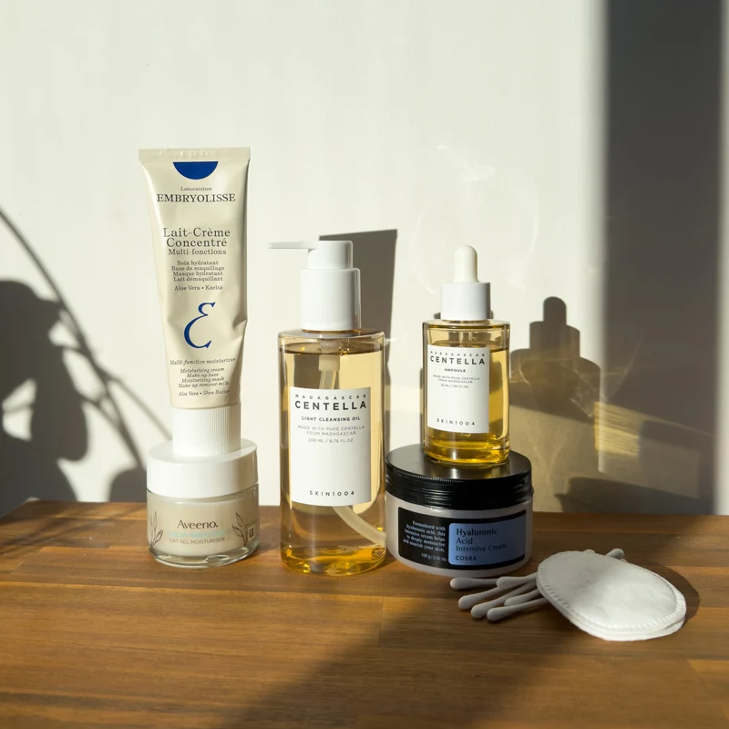 Skincare products on a wooden surface including Embryolisse cream, Skin1004 cleansing oil, serum, Aveeno gel, and COSRX cream.