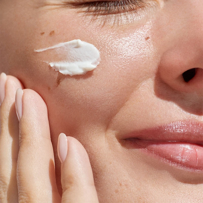 Close-up of fingers applying white facial moisturizer cream to cheek, showing smooth skin texture and natural lips.