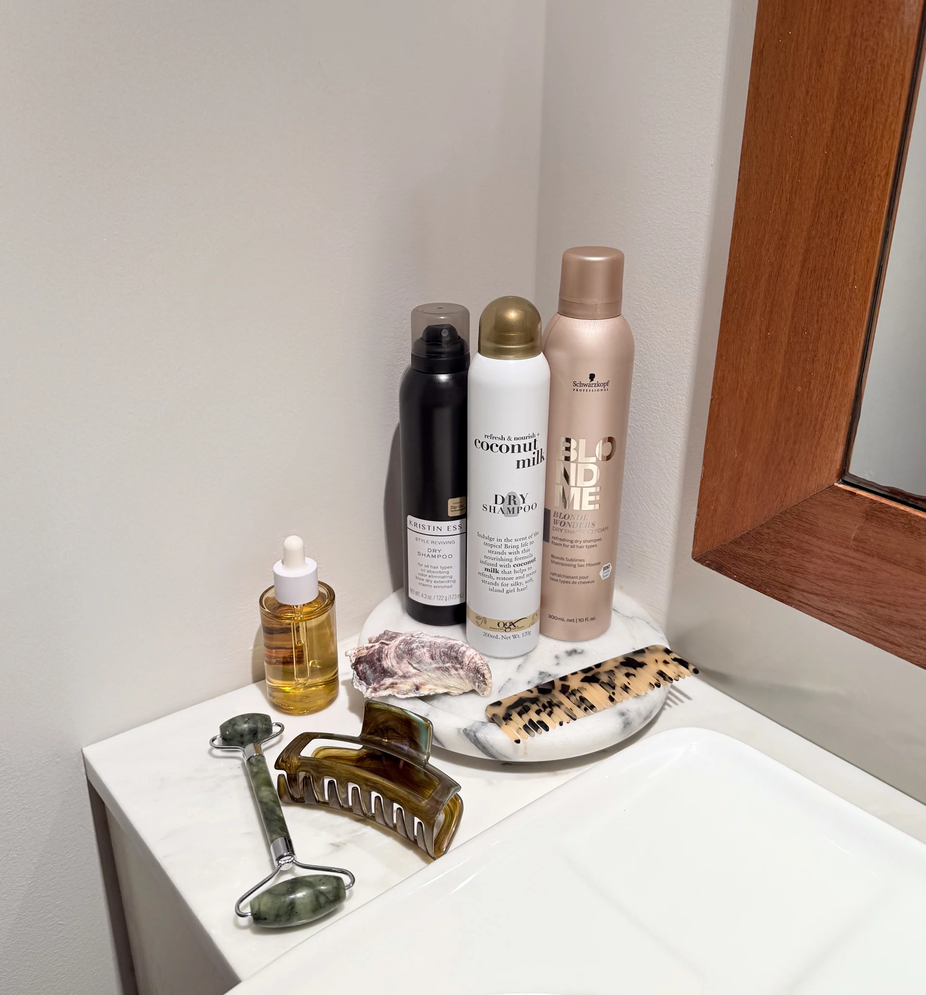 Hair care products including dry shampoo, styling sprays, and facial roller displayed on white surface with marble accessories.