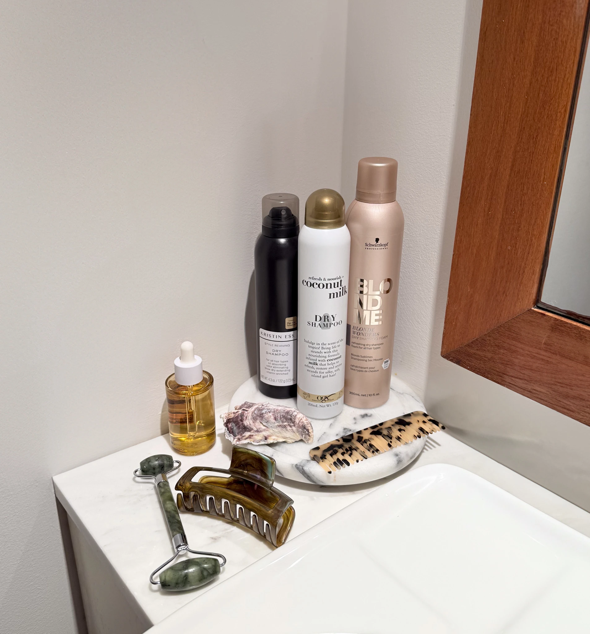Hair care products including dry shampoo, styling sprays, and facial roller displayed on white surface with marble accessories.