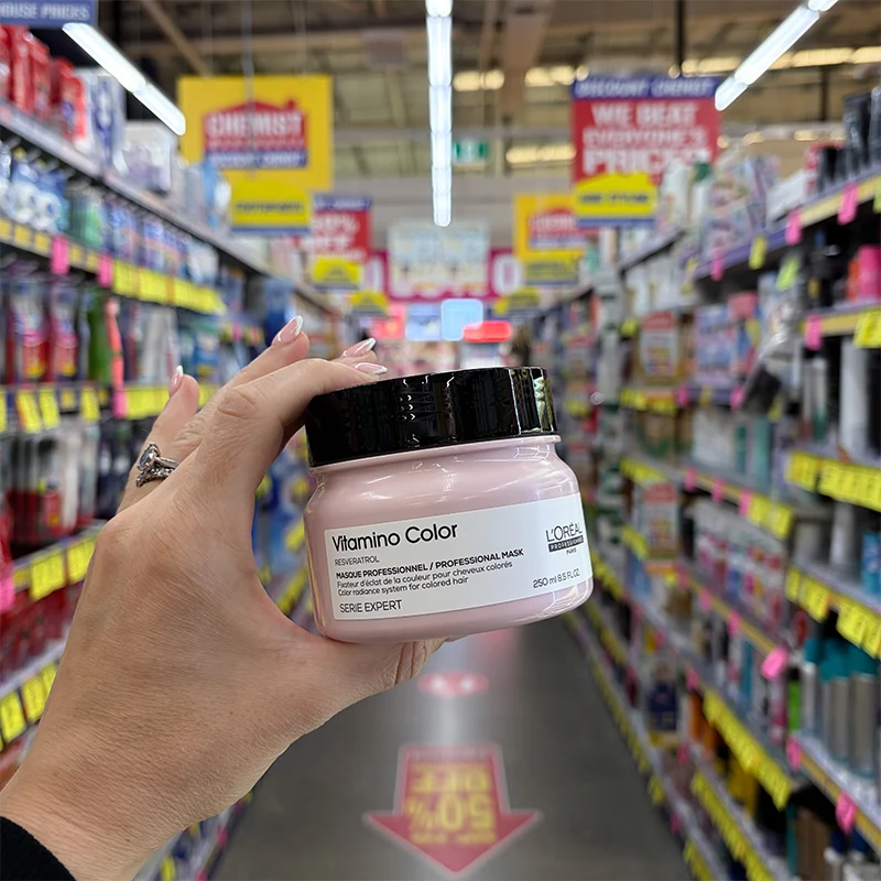 A hand holding a pink jar of L'Oreal Serie Expert Vitamino Color Professional Mask in a pharmacy store aisle.