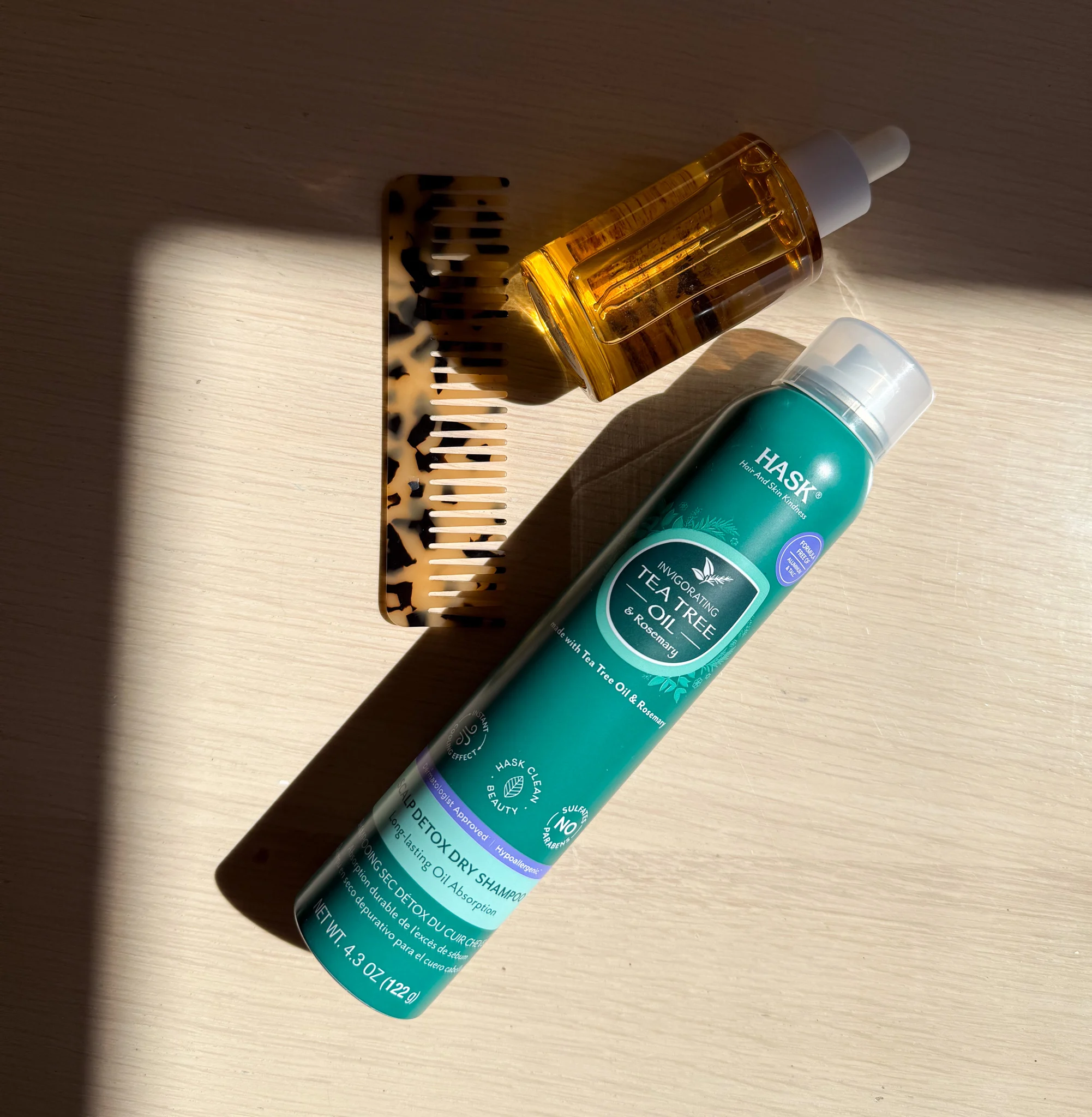 Hair care products including a turquoise dry shampoo bottle, amber-colored oil dropper, and tortoiseshell comb on light wooden surface.
