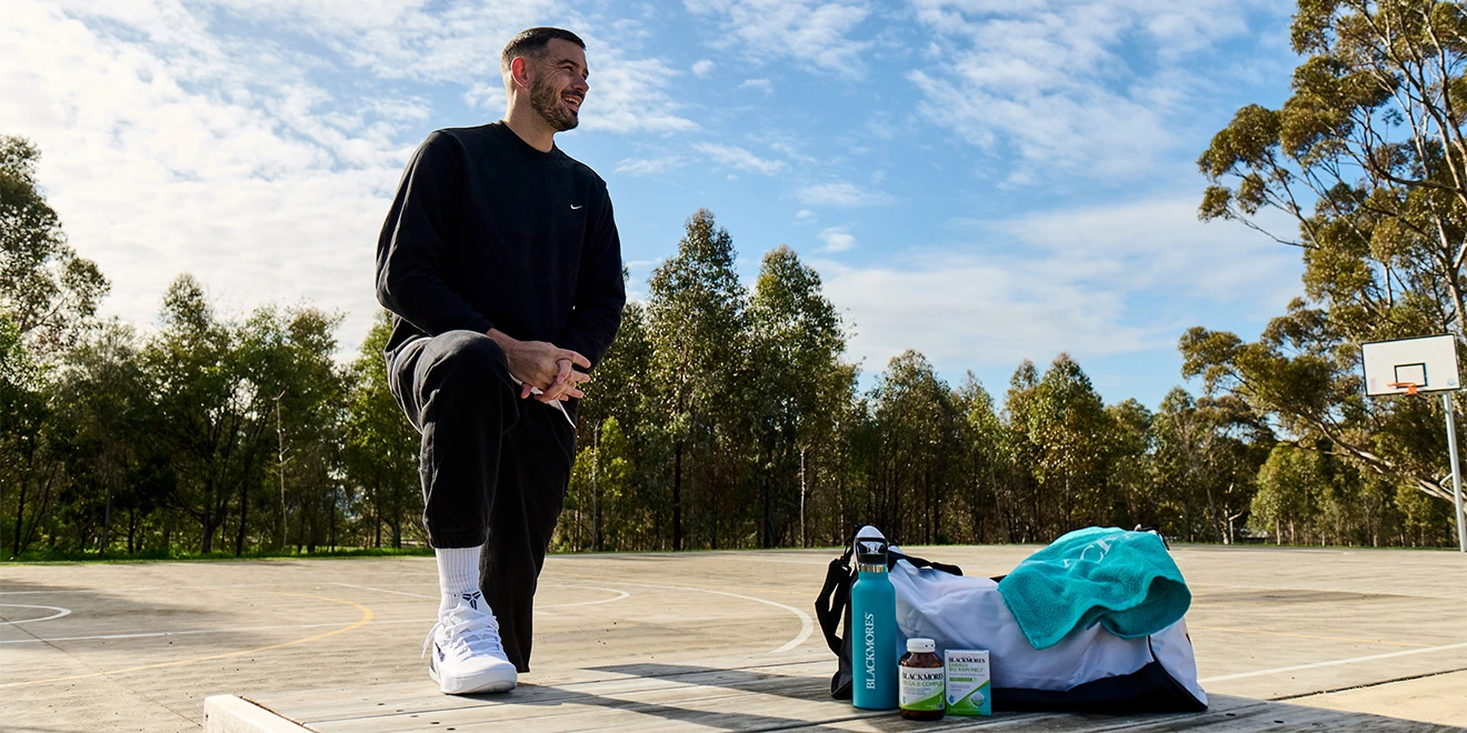 Person in black Nike sportswear standing on outdoor basketball court with gym bag and sports equipment against tree backdrop.