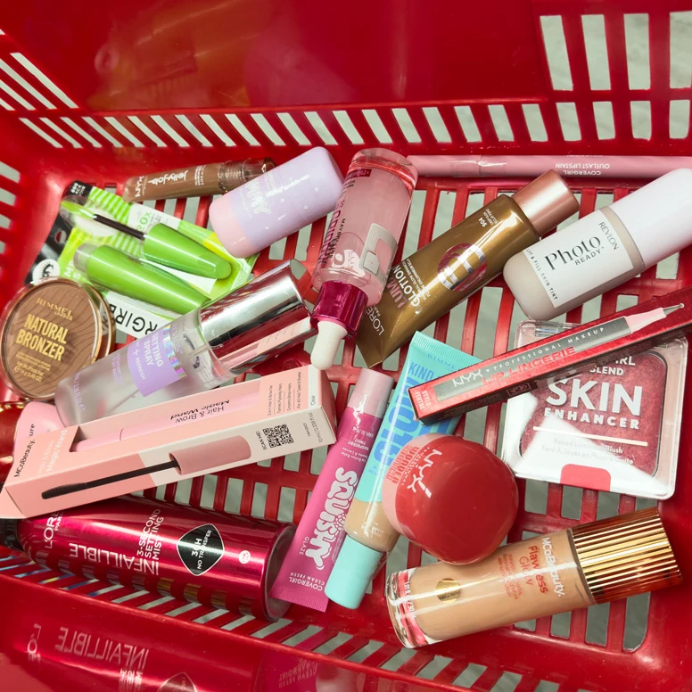 A red shopping basket filled with various makeup products including foundations, bronzer, lip gloss, and blush from brands like NYX, L'Oreal, and Revlon.