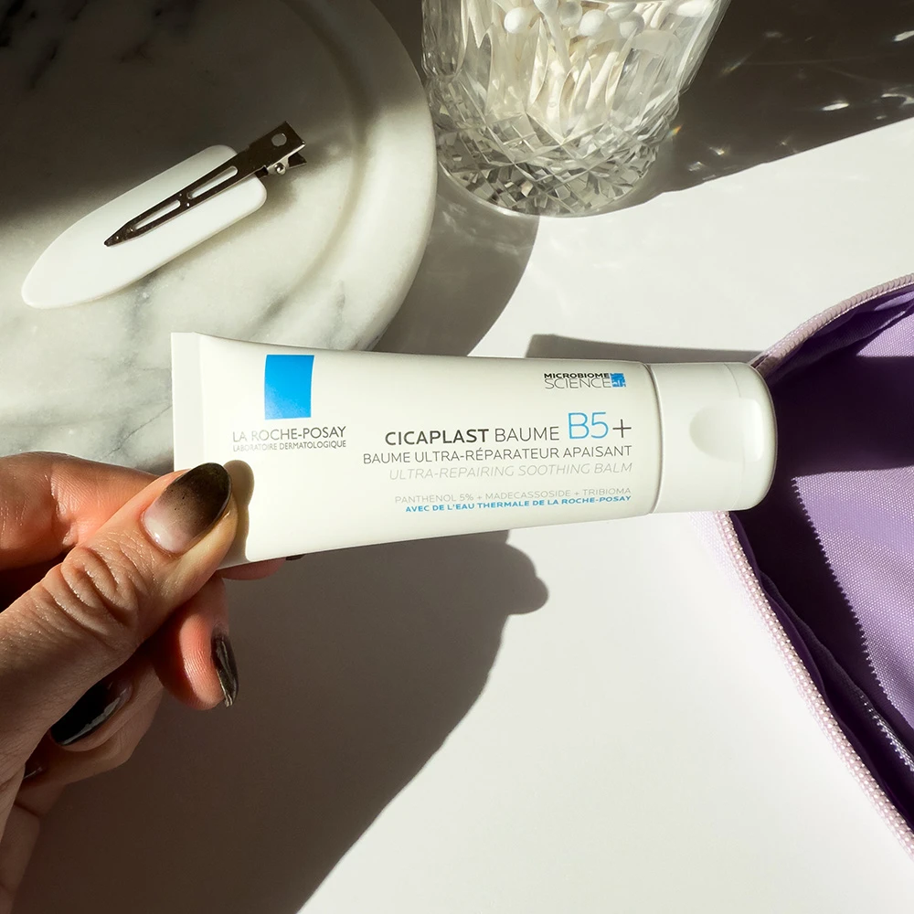 A tube of La Roche-Posay Cicaplast Baume B5+ cream held against a white surface with a crystal glass and purple fabric visible