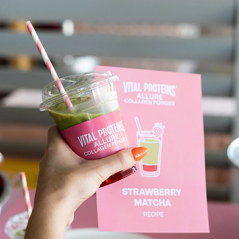 Hand holding green matcha drink in plastic cup with pink Vital Proteins Collagen Powder packaging in background.