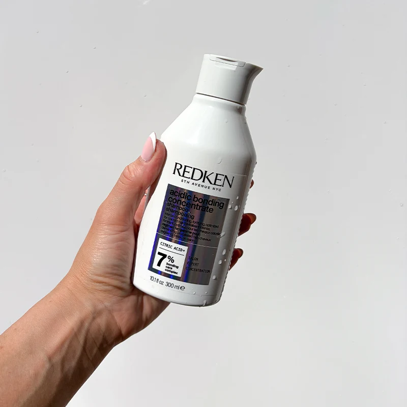 Hand holding a white Redken hair product bottle with purple label details against a light background.