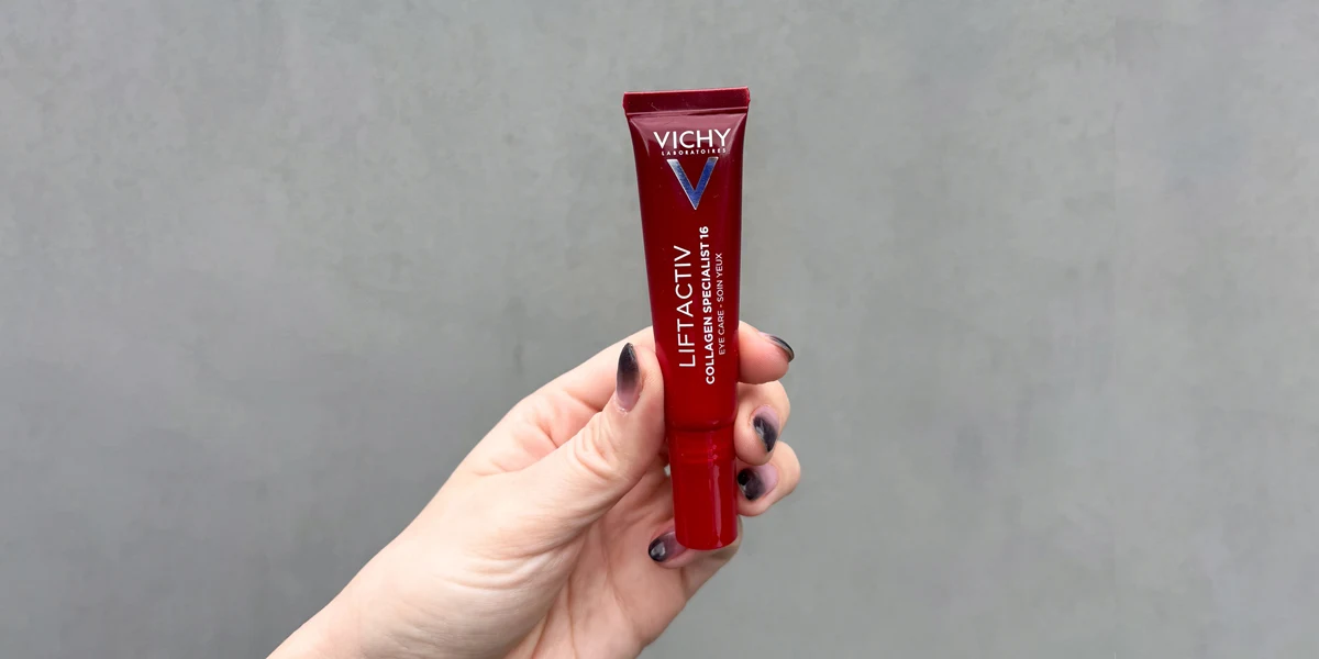 A hand with dark nails holds a red tube of Vichy Liftactiv Collagen Specialist 16 Eye Care against a grey background.