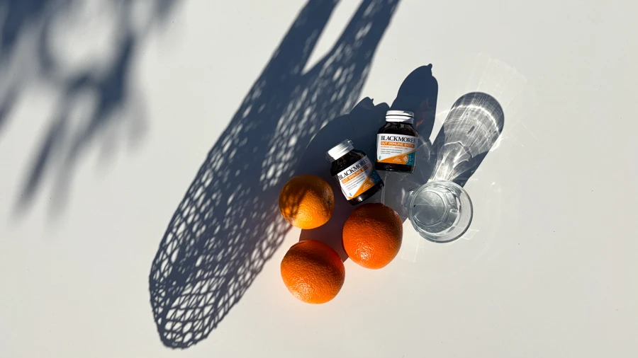 Two Blackmores supplement bottles, three oranges, and a glass of water on a white surface with net bag shadow.