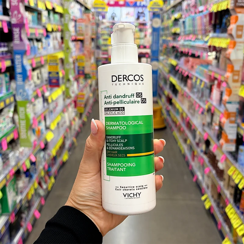 A hand holding a white bottle of Vichy Dercos Anti-Dandruff Dermatological Shampoo in a store aisle.