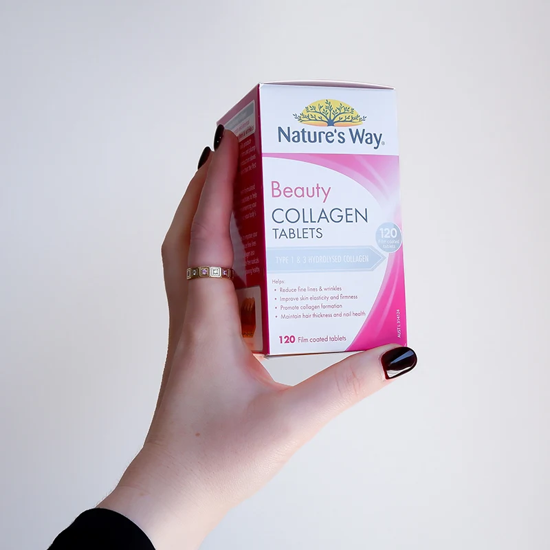 Hand with black nail polish holding Nature's Way Beauty Collagen supplement box containing 120 tablets against light grey background.