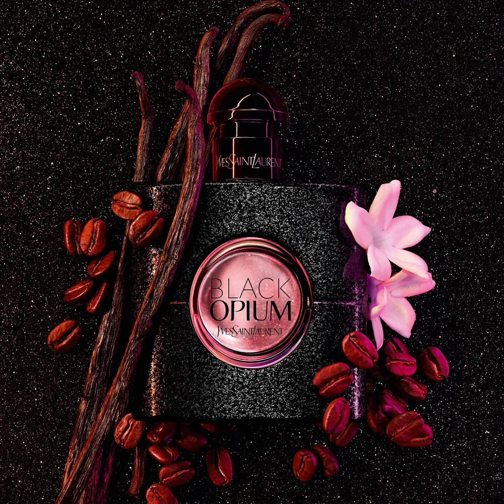YSL Black Opium perfume bottle with vanilla beans, coffee beans, and pink flowers on dark glittery background