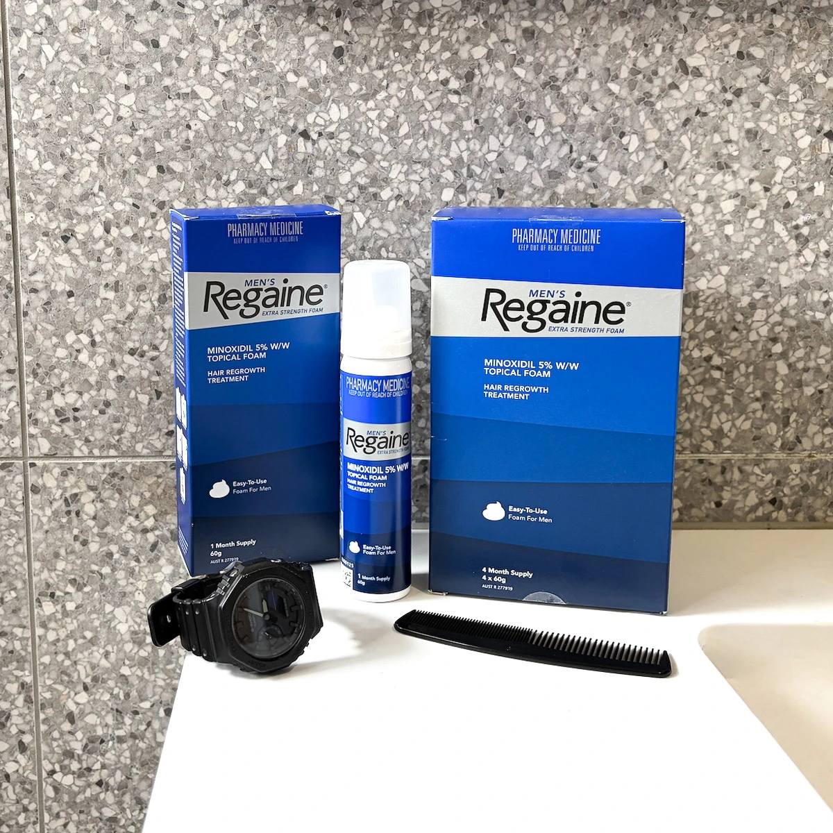 Two blue Regaine Men's Extra Strength Foam boxes, a foam canister, a black comb, and a watch on a bathroom counter.