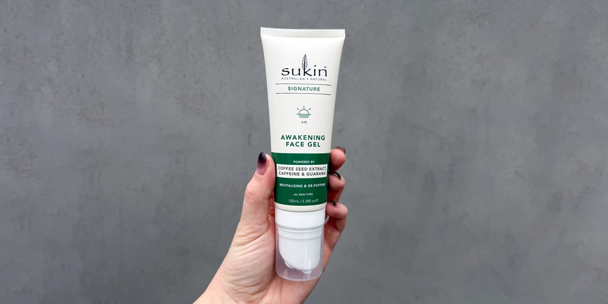 A hand holding a white tube of Sukin Signature Awakening Face Gel with green label, against a grey background.