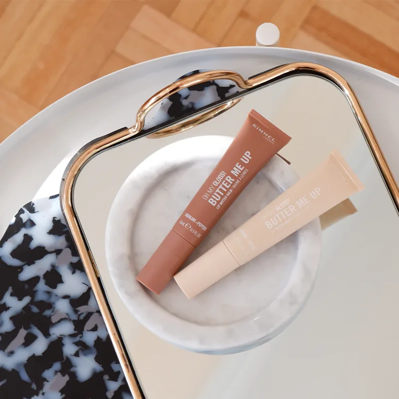 Two Rimmel Butter Me Up lip products in beige and brown tubes on white ceramic dish with gold-rimmed decorative mirror.