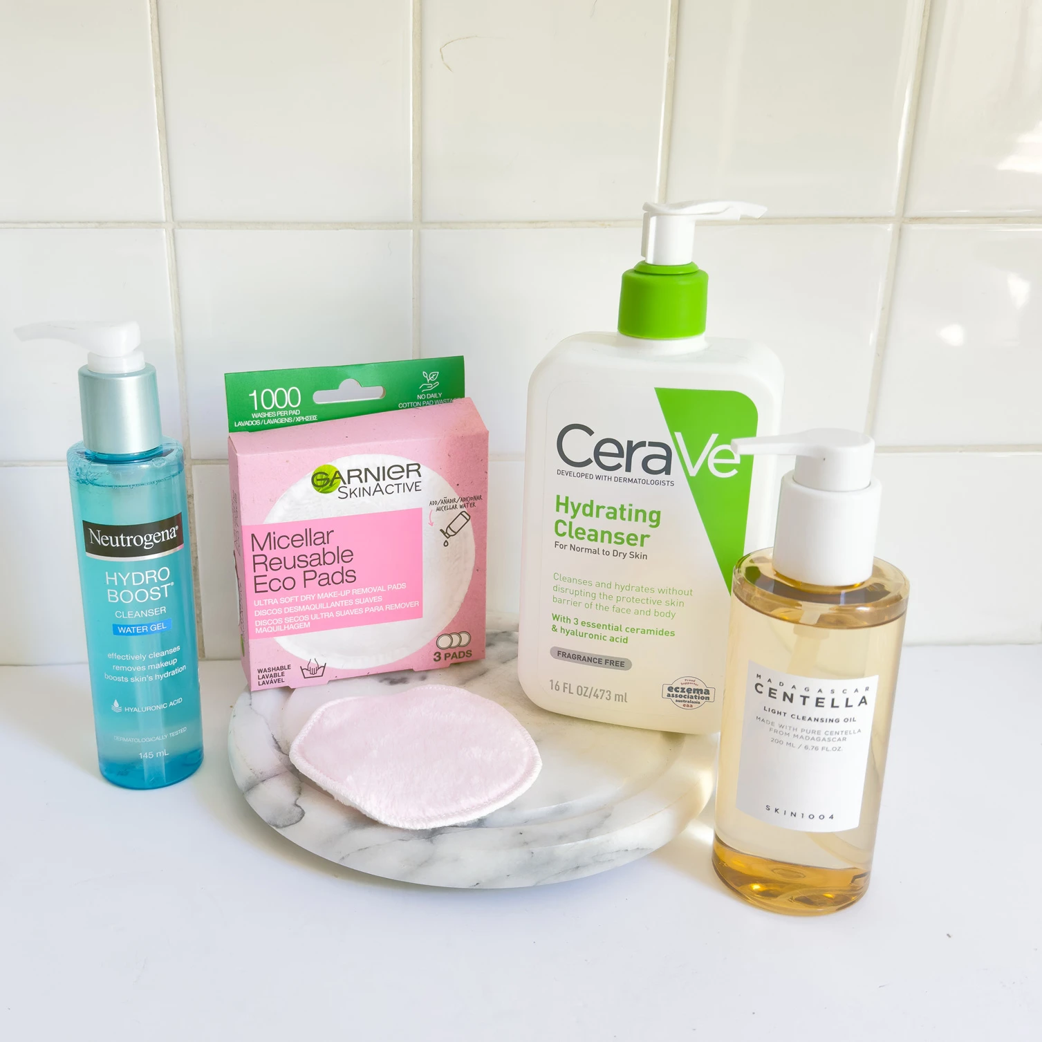 Collection of skincare products including Neutrogena, CeraVe, and Garnier cleansers with reusable cotton pads on marble surface