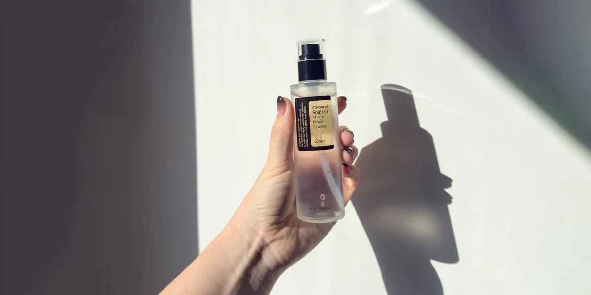 A hand holding a COSRX Advanced Snail 96 Mucin Power Essence bottle against a white background with soft shadows.