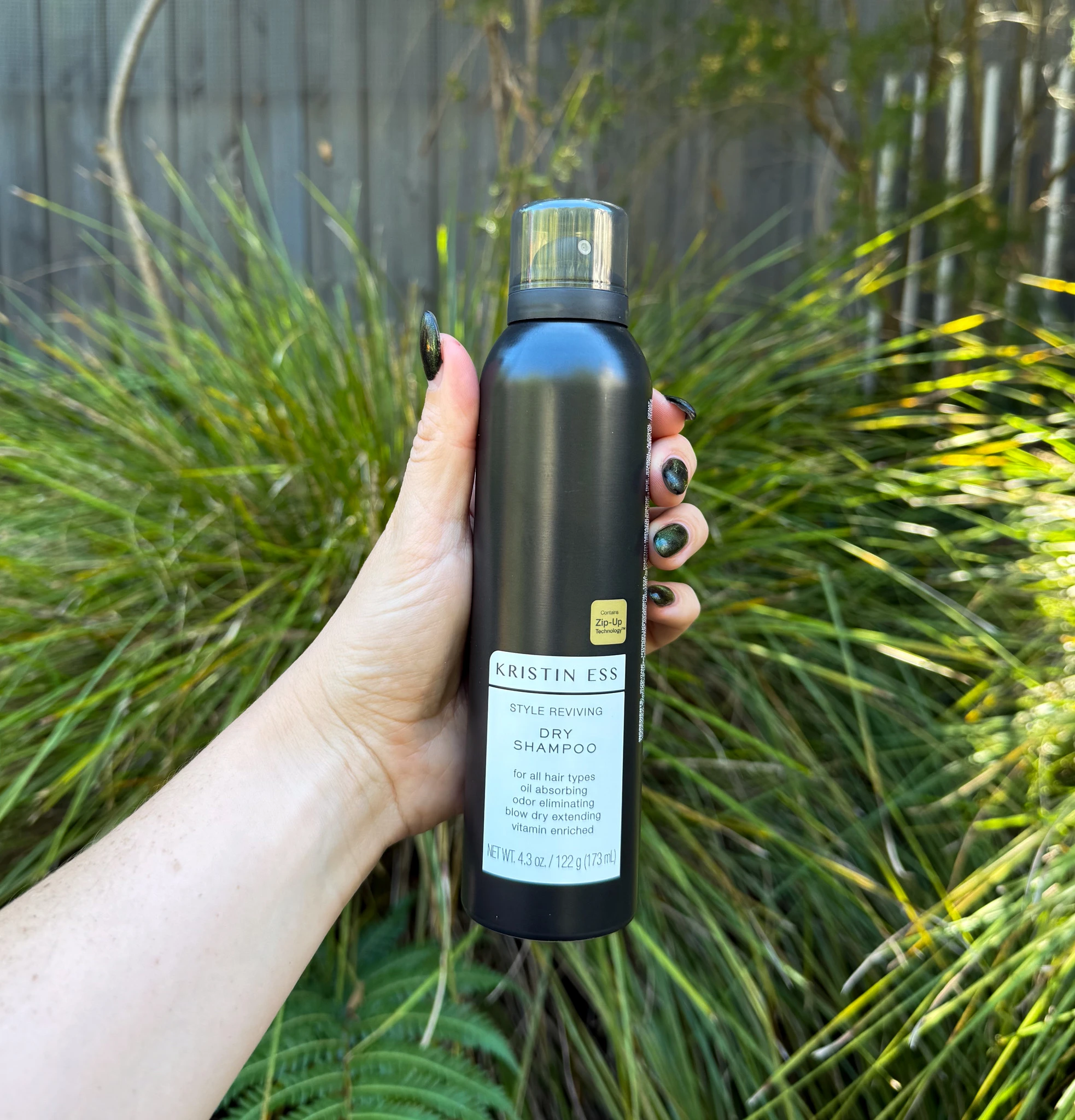 Hand holding black Kristin Ess dry shampoo bottle against green grass background, showing dark green nail polish.