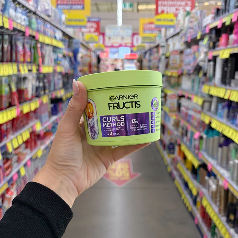 A hand holds a green tub of Garnier Fructis Curls Method Step 3 Mask in a store aisle with shelves of hair products.