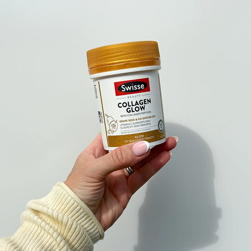 Hand in cream sweater holding a bottle of Swisse Collagen Glow supplements with gold cap against white background