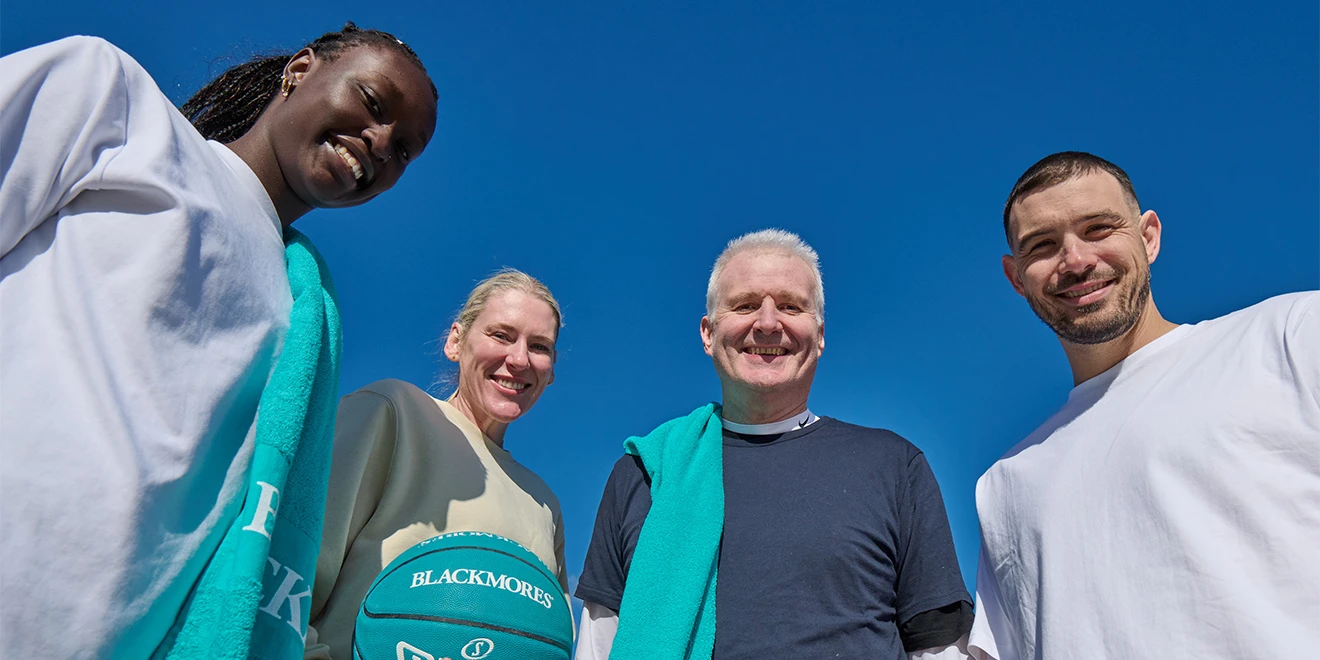 Four smiling people in casual sportswear pose with a turquoise basketball against a bright blue sky, photographed from below.