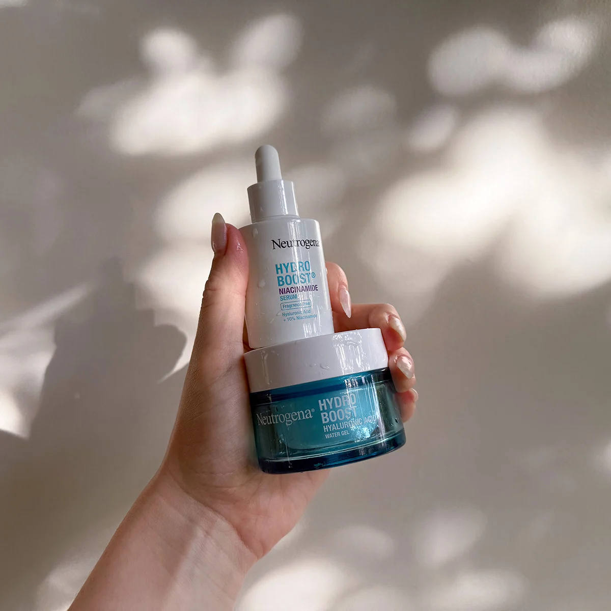 Hand holding Neutrogena Hydro Boost skincare products - a white serum dropper and blue gel moisturiser - against a shadowy background.