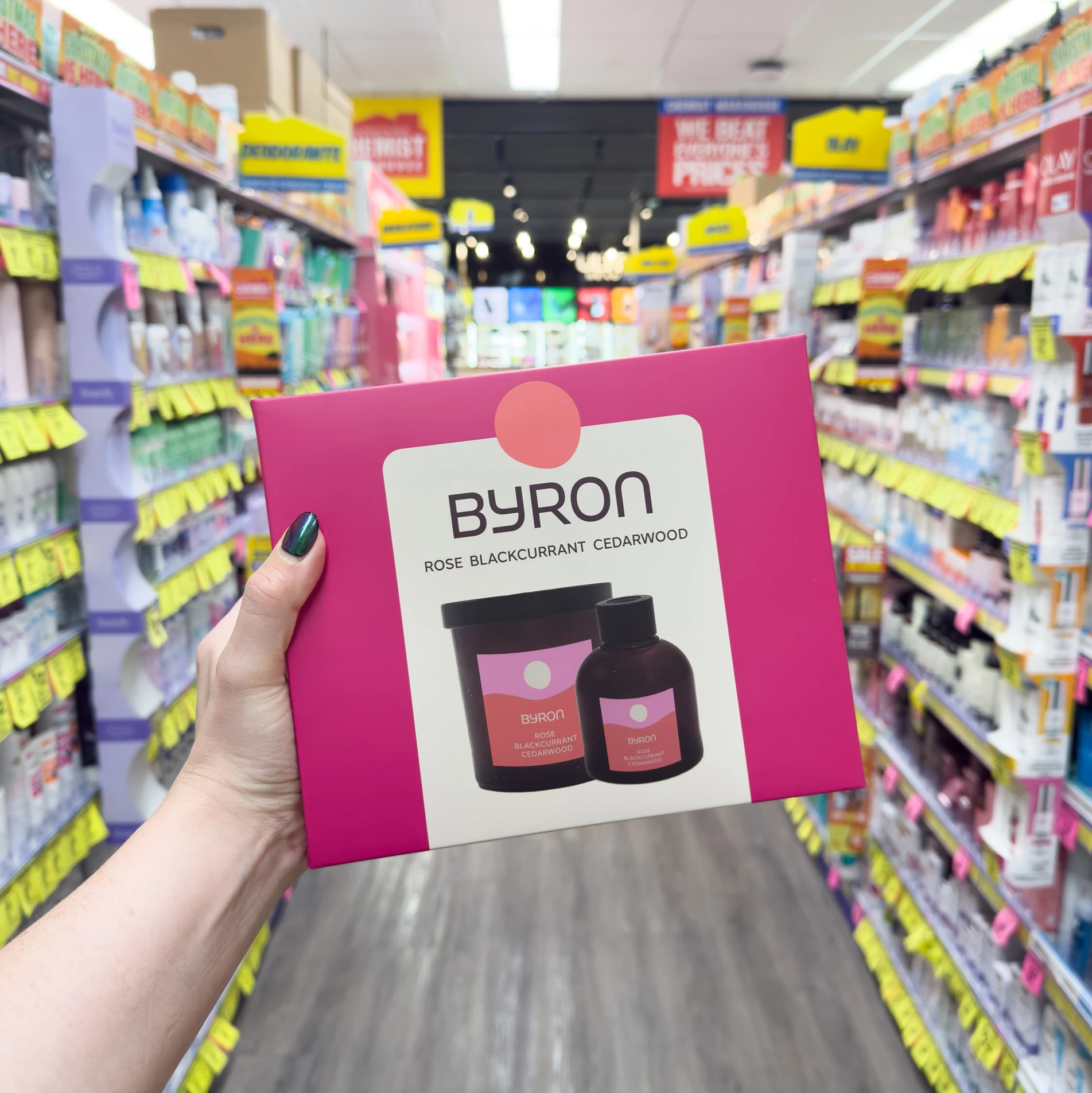 Hand holding Byron fragrance card showing rose blackcurrant cedarwood perfume bottles against backdrop of retail store shelves