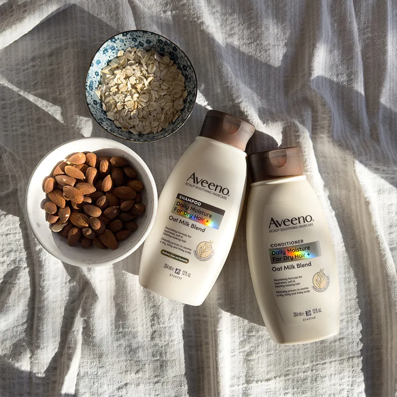 Aveeno shampoo and conditioner bottles with oat milk blend, alongside bowls of almonds and oats on white fabric in natural light.