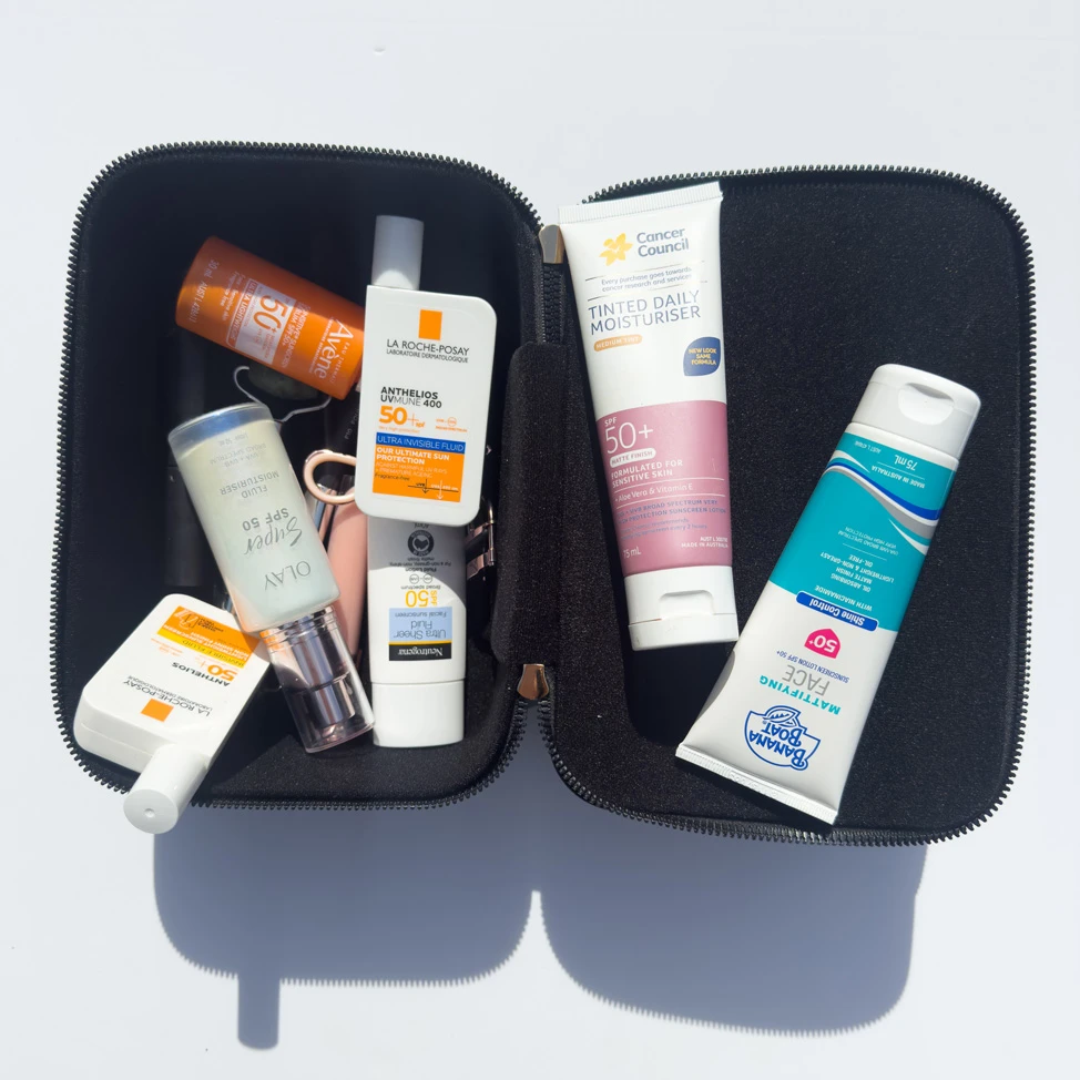 Open black toiletry bag containing multiple SPF50+ sunscreen products including La Roche-Posay, Cancer Council, Banana Boat, Olay, and Neutrogena.