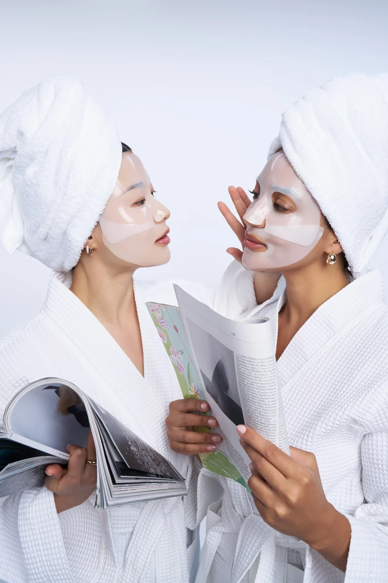 Two people in white bathrobes and towels wearing face masks while reading magazines during a spa treatment