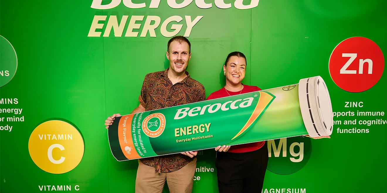 Two people holding large Berocca Energy prop tube against green backdrop with vitamin symbols and branding elements.