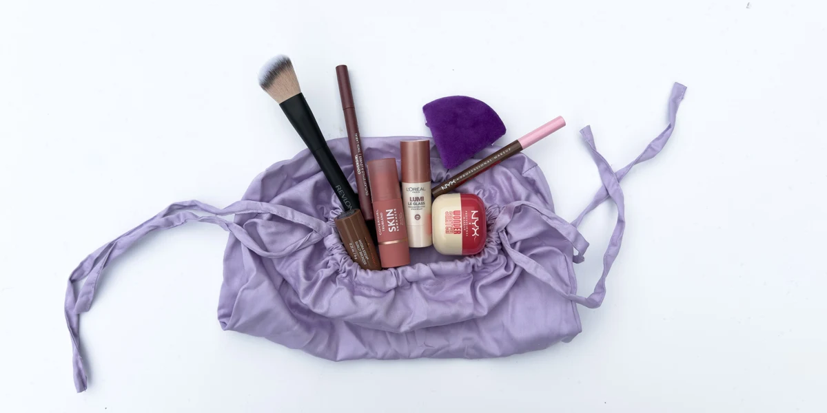 A lavender drawstring makeup bag open and filled with various cosmetics including brushes, lip products, and NYX foundation.
