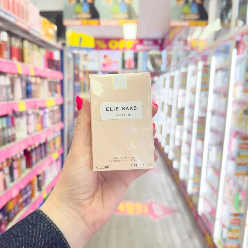 A hand with red nails holds an Elie Saab Le Parfum 30ml box in a store aisle with colorful shelves in the background.