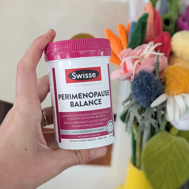 A hand holding a white bottle with a pink lid of Swisse Ultiboost Perimenopause Balance tablets, with colorful flowers in the background.