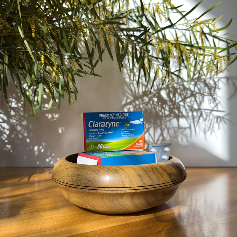 Box of Claratyne allergy medication resting in a wooden bowl on a table, with wattle branches casting shadows above