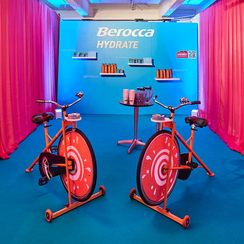 Orange exercise bikes with red spiral wheel designs in a blue room with pink curtains and Berocca Hydrate branding on the wall.