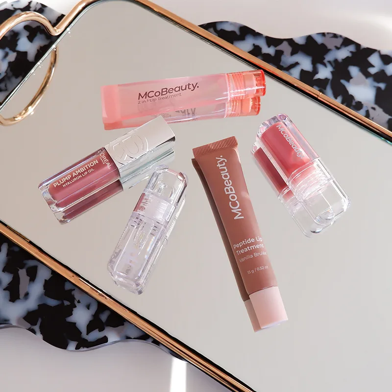 Collection of MCoBeauty lip products including glosses and treatments displayed on reflective surface with tortoiseshell pattern border.