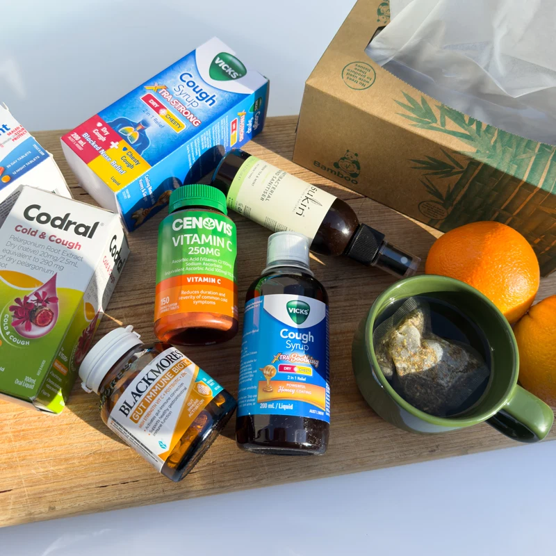Cold and flu remedies on a wooden board including Vicks cough syrup, Codral, Cenovis Vitamin C, Blackmores, tea, and oranges.