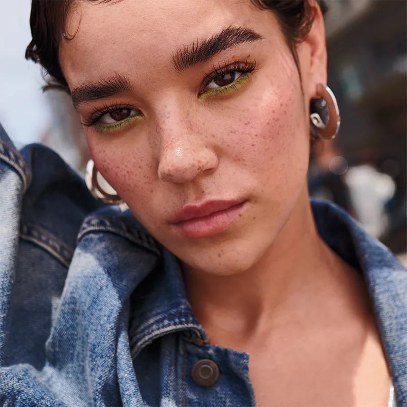 Close-up portrait showing bright lime green eyeshadow, natural freckles, and silver hoop earrings, wearing a denim jacket.