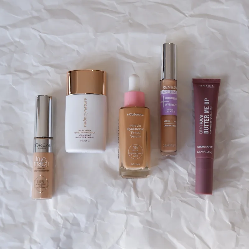 Five makeup products including foundations, serums, and concealer from L'Oreal, Revlon, and other brands on white crumpled paper background.