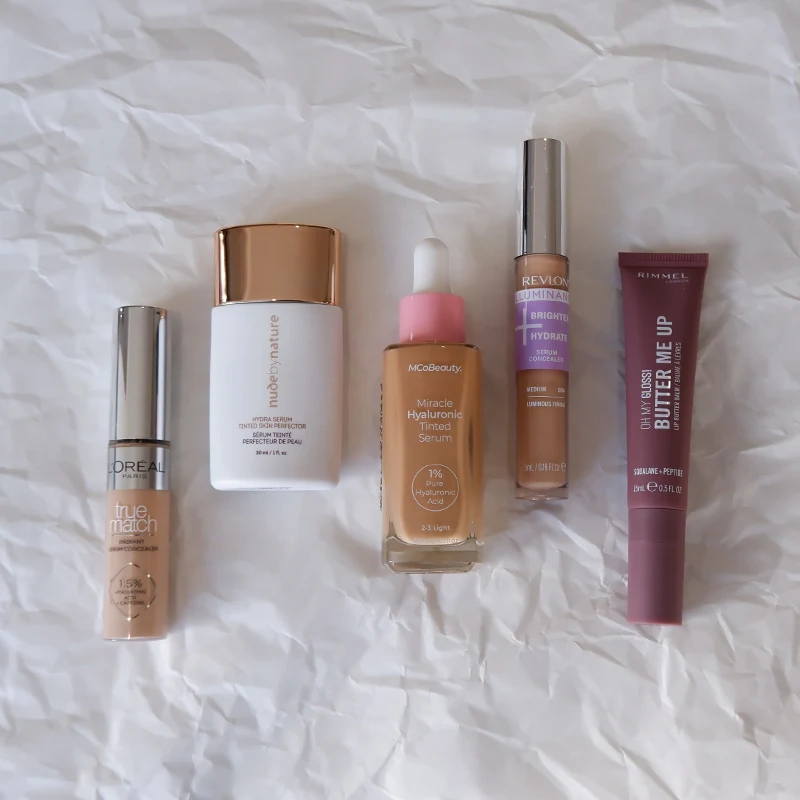 Five makeup products including foundations, serums, and concealer from L'Oreal, Revlon, and other brands on white crumpled paper background.