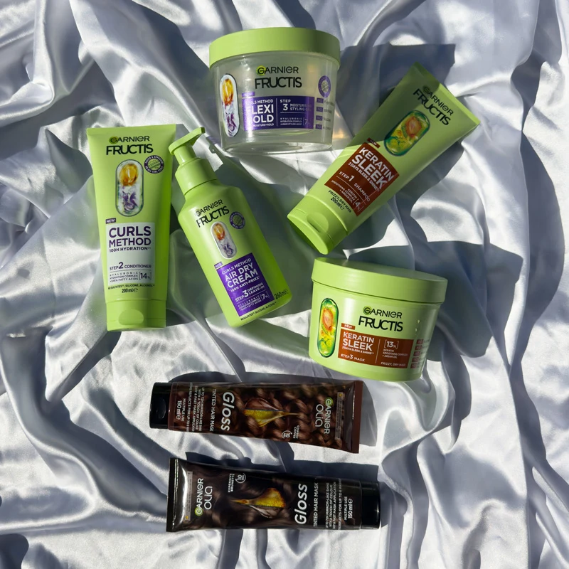 Garnier Fructis hair care products including Curls Method, Keratin Sleek, and Olia Gloss arranged on white satin fabric.