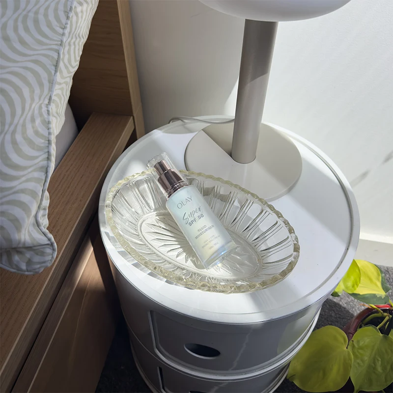 Olay skincare product displayed on decorative glass dish atop white modern side table with green plant leaves visible below.