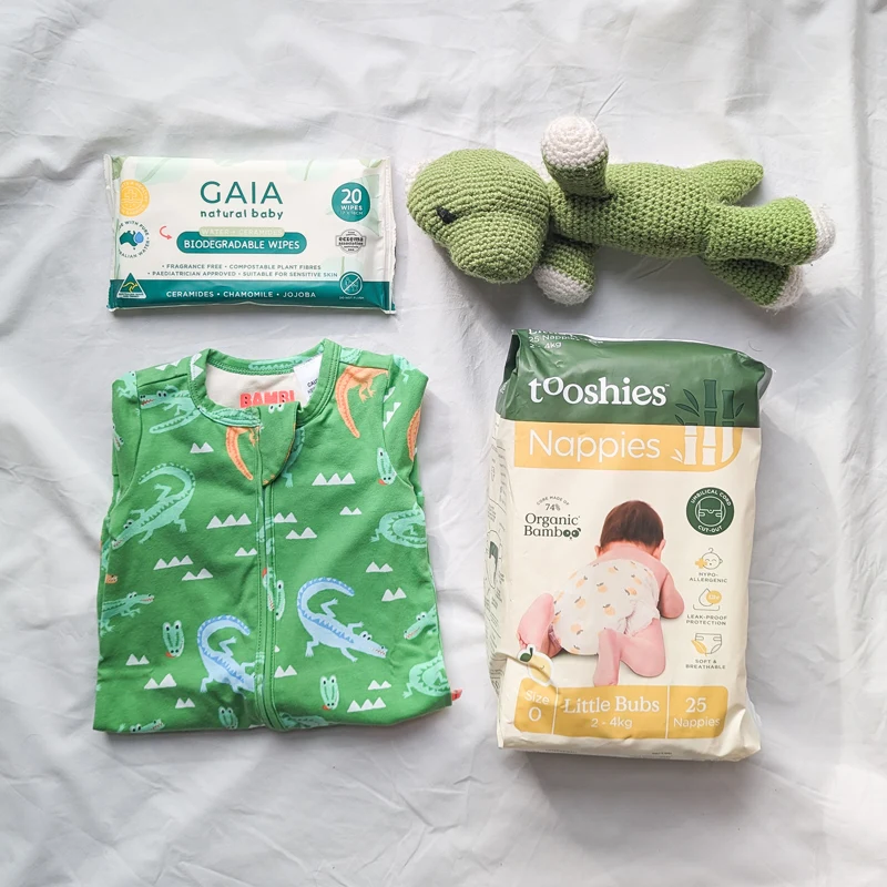 Baby essentials flat lay: GAIA biodegradable wipes, green crochet dinosaur toy, crocodile print onesie, and Tooshies bamboo nappies.