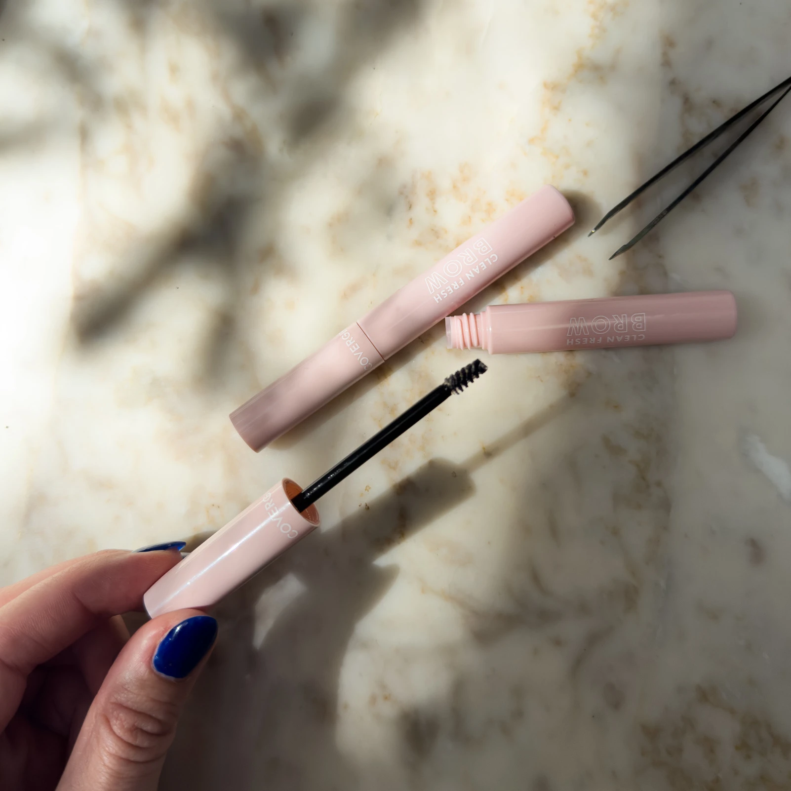 Pink eyebrow gel tubes on marble surface, with applicator brush being held by hand with blue nail polish
