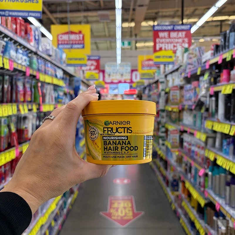 A hand holding a yellow tub of Garnier Fructis Banana Hair Food mask in a chemist store aisle.