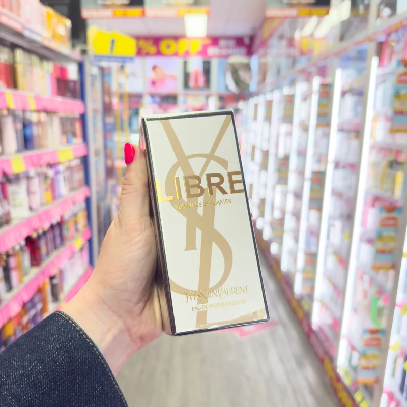 Hand with red nails holding YSL Libre Flowers & Flames Eau de Parfum box in a fragrance store aisle.