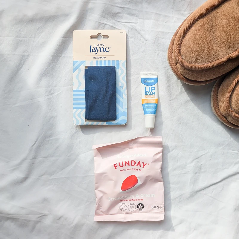 Lady Jayne blue headband, Dermal lip balm tube, Funday strawberry gummies pack, and tan suede slippers on a white fabric surface.