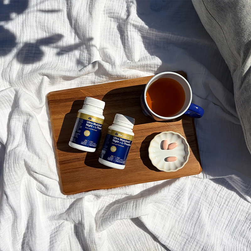 Two BioCeuticals Ultra Muscleze Night + L-Theanine supplement bottles on a wooden tray with tea and pink tablets.
